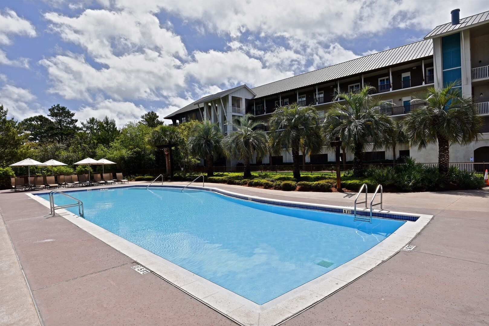 Una piscina rectangular con una escalera metálica, rodeada de sillas de descanso y sombrillas, con un edificio al fondo.