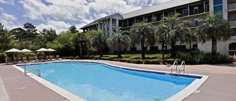 Una piscina rectangular con una escalera metálica, rodeada de sillas de descanso y sombrillas, con un edificio al fondo.