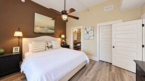 A bedroom with a bed, nightstands, a ceiling fan, a door, and a wall with a painting. - Vitamin Sea - 3BR/3BA Redfish Village Condo - Sleeps 9 - Deeded Beach Access (Santa Rosa Beach)