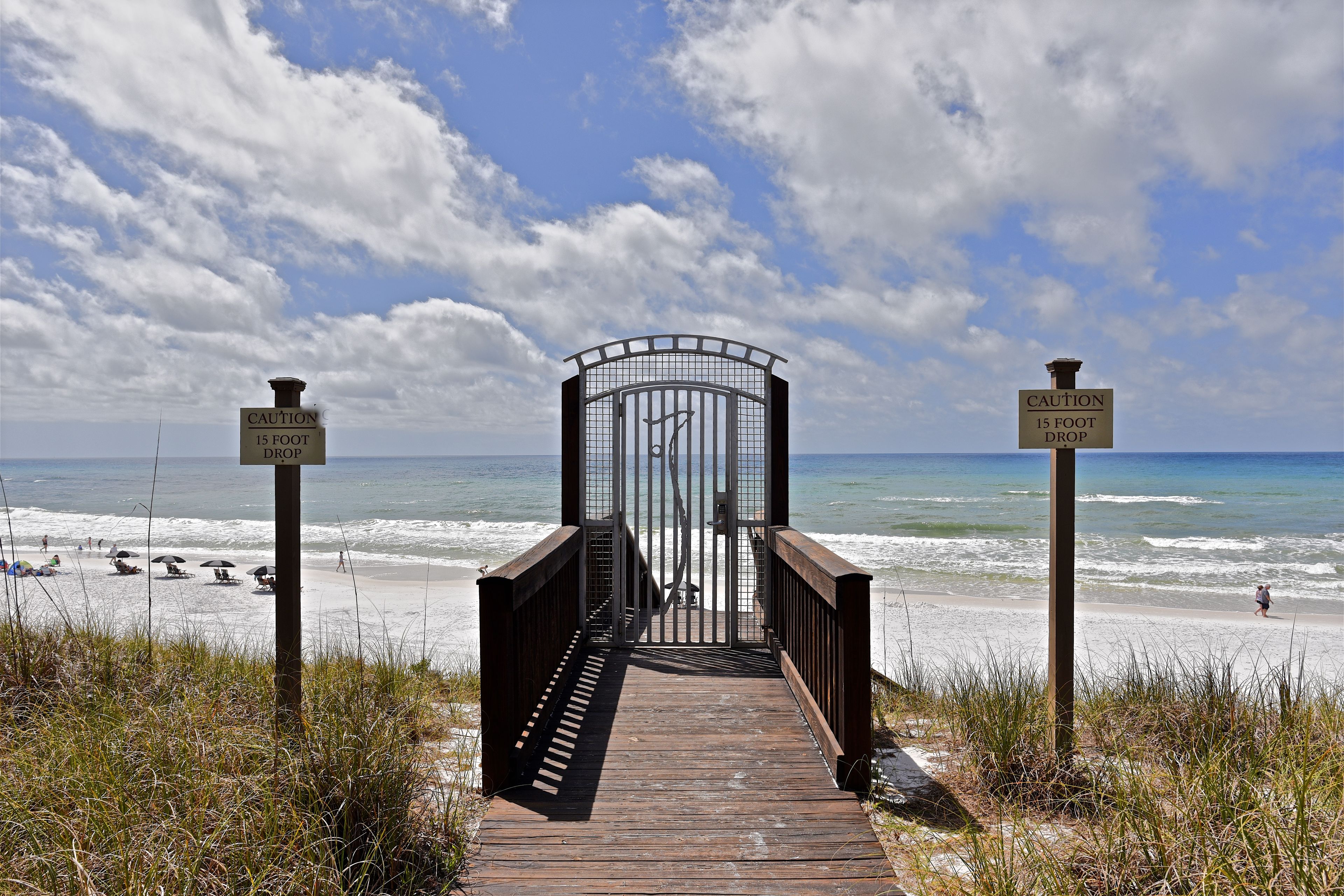 A wooden boardwalk leading to a beach with caution signs indicating a 15-foot drop.