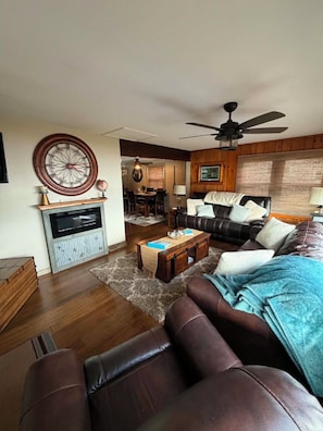 A living room with a fireplace, a ceiling fan, a dining area, and a view of the outdoors.