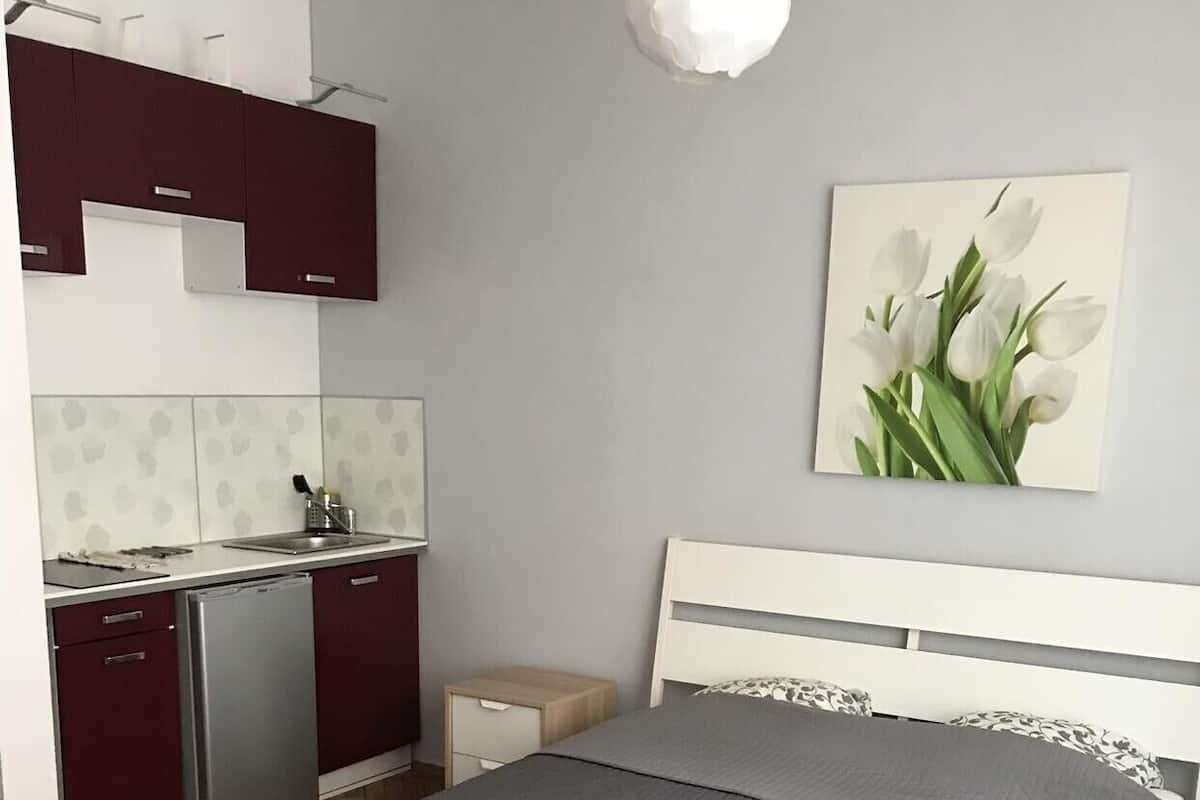 Quarto com cama, mesa de cabeceira, escrivaninha, geladeira e cozinha equipada com armários e pia.