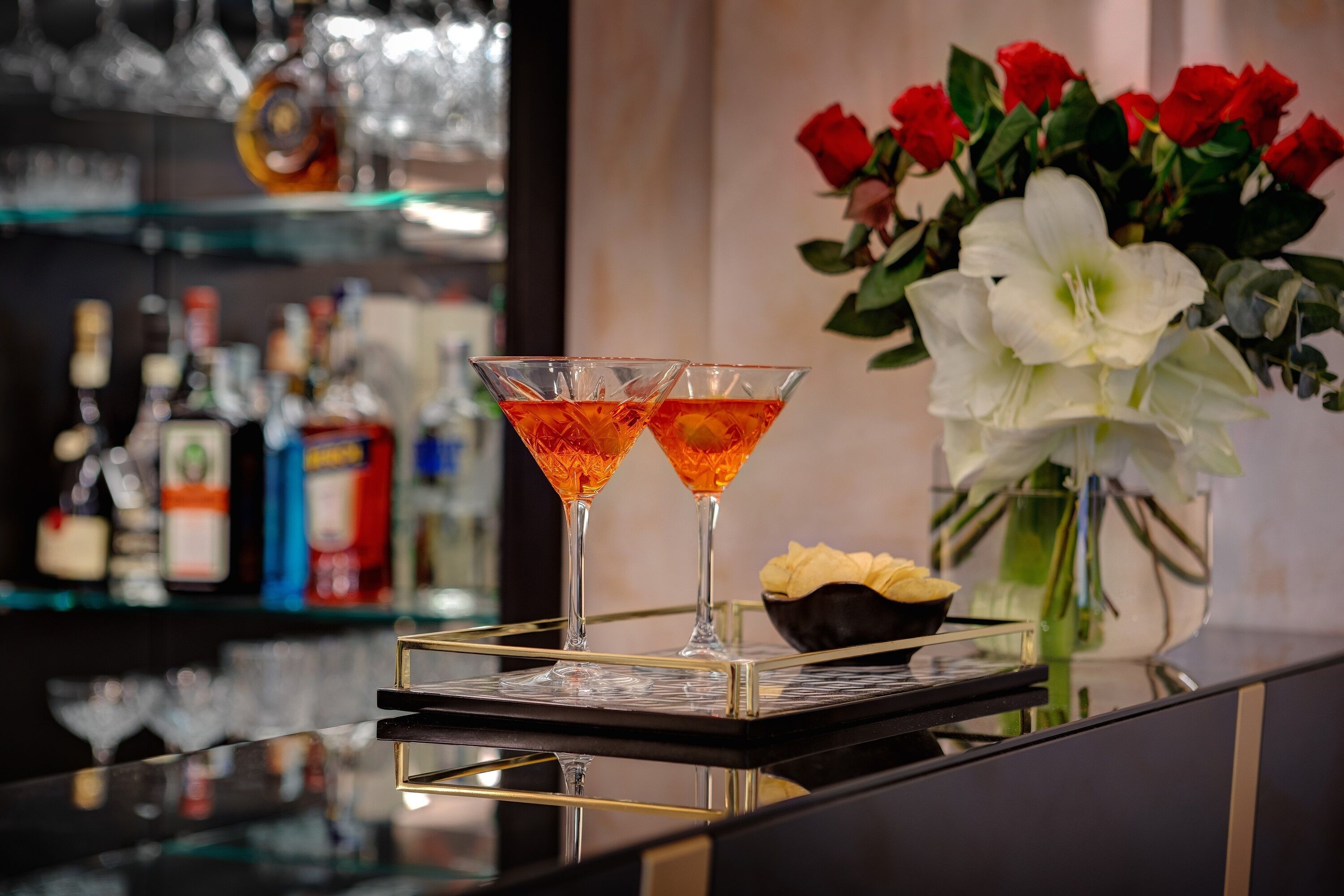 A bar area with two martini glasses on a tray, a vase with red and white flowers, and a bowl of snacks.