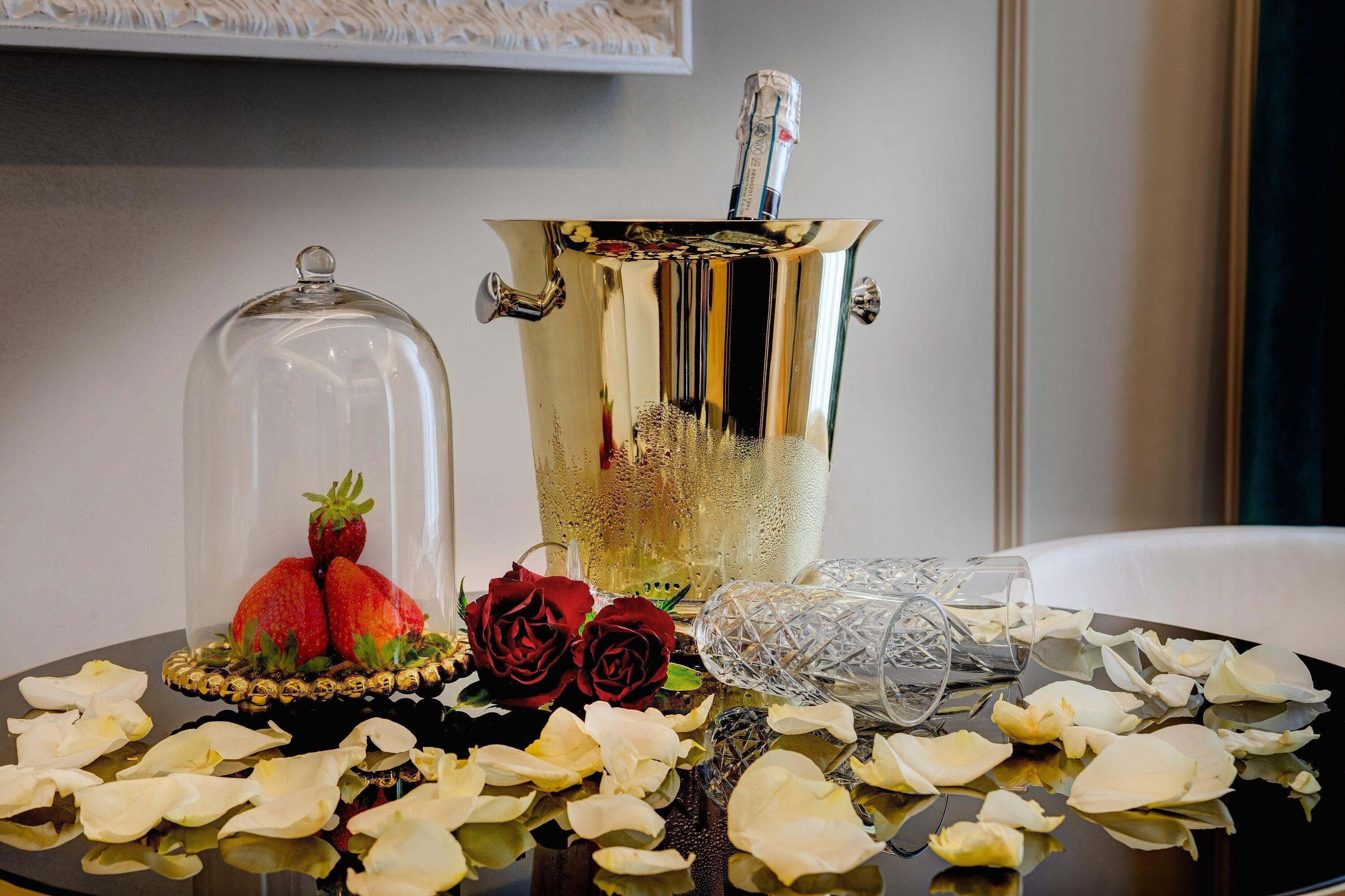Ein Tisch mit einem Champagnerkühlereimer, einer Glaskuppel mit Erdbeeren, zwei Rosen und verstreuten Blütenblättern.