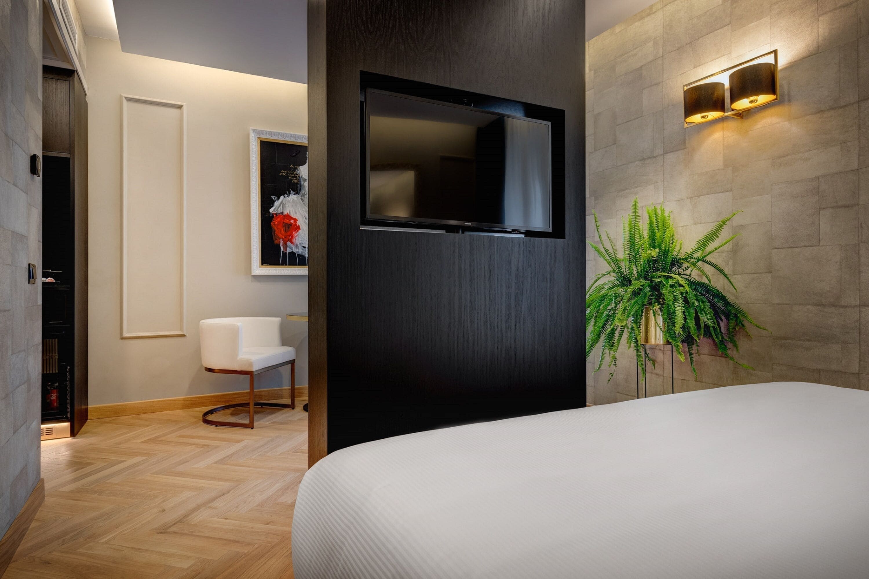 A modern hotel room with a large bed, a flat-screen TV mounted on the wall, a white chair, and a potted plant.