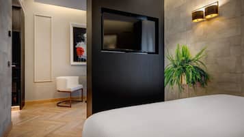 A modern hotel room with a large bed, a flat-screen TV mounted on the wall, a white chair, and a potted plant.