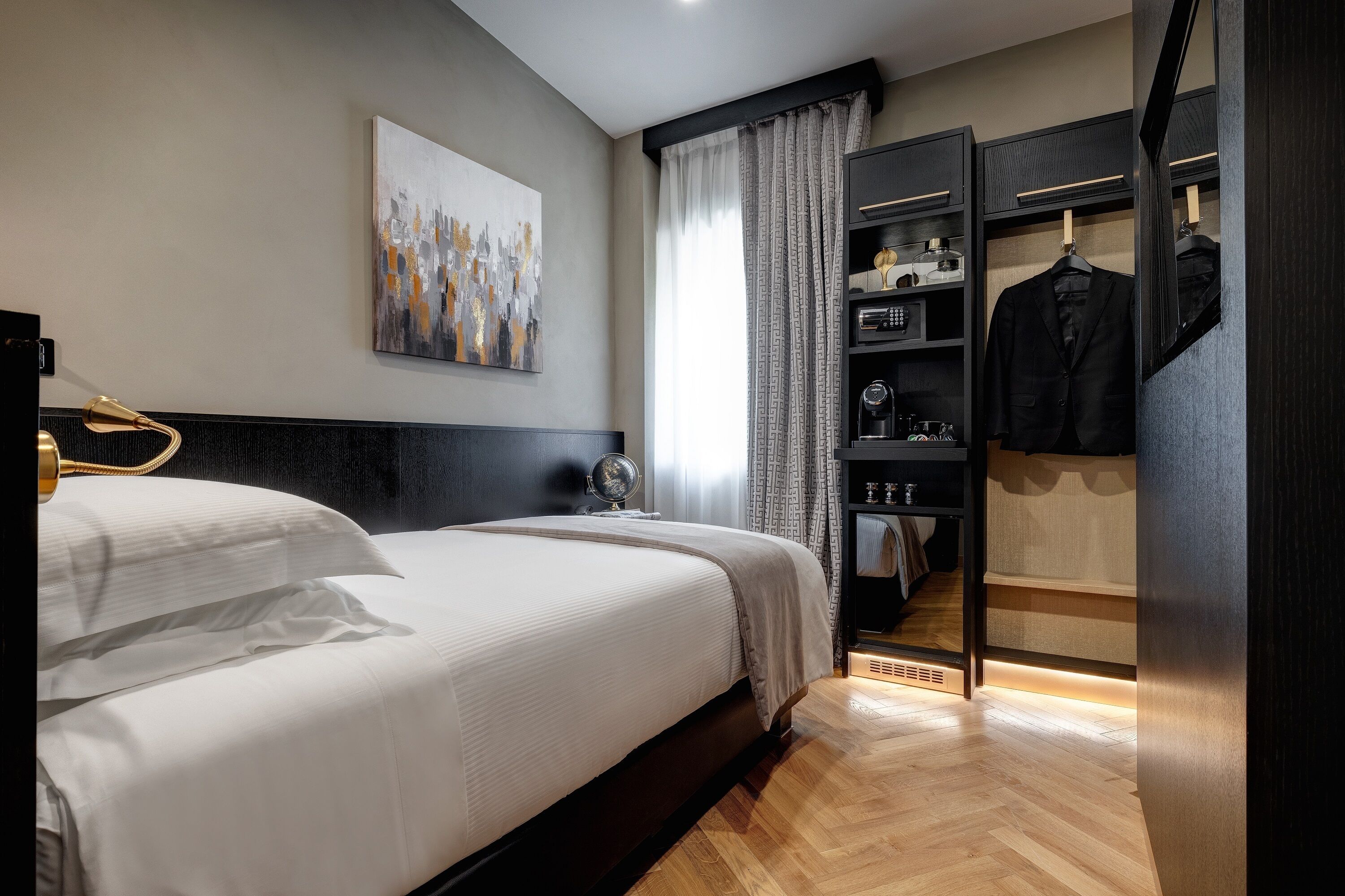 Une chambre d’hôtel moderne avec un lit bien fait, une table de chevet avec une lampe, une armoire où sont suspendus des vêtements et un tableau accroché au mur.