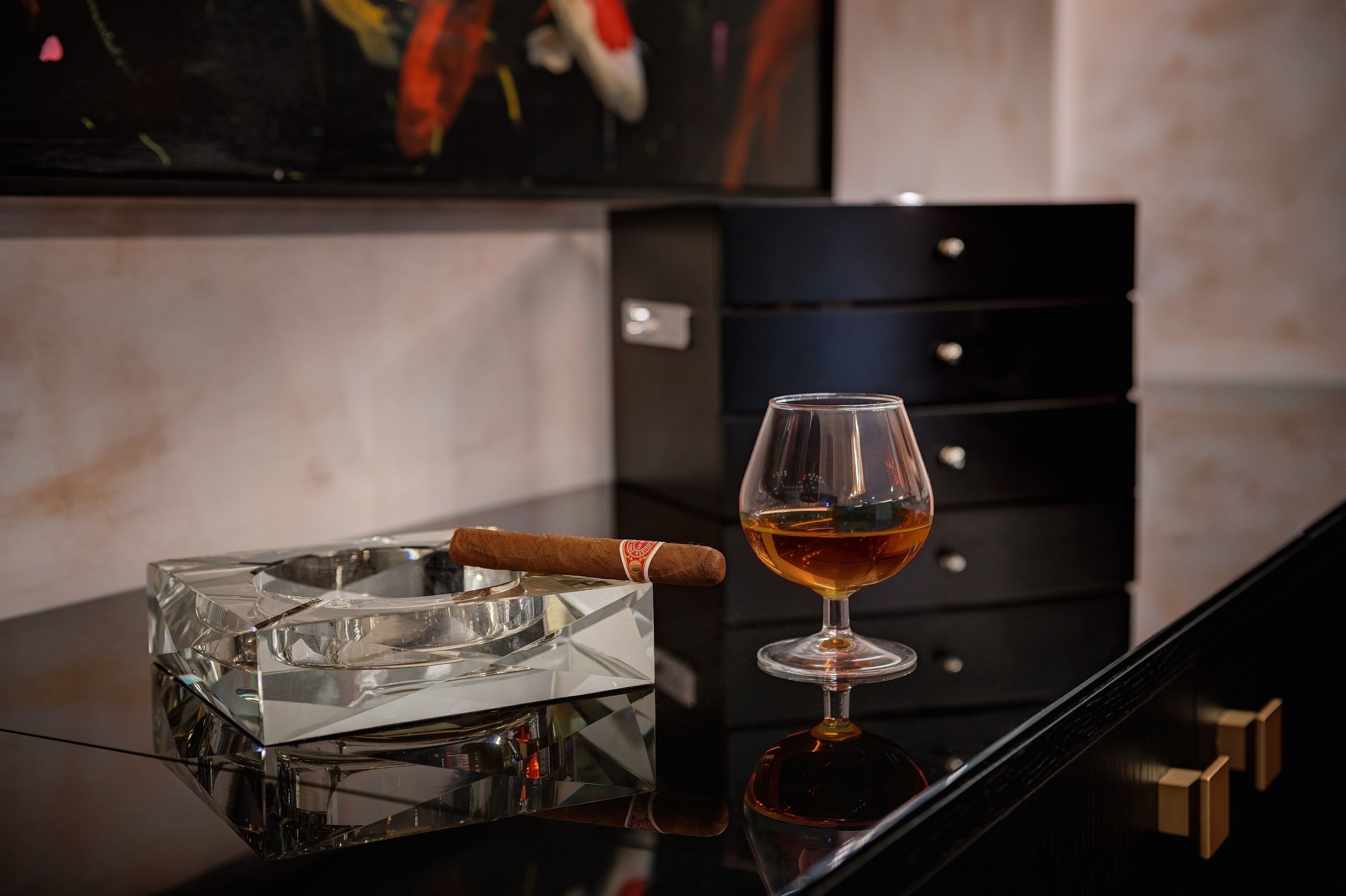 A glass ashtray with a cigar on a reflective surface, a glass of amber liquid, and a black drawer unit with gold handles.