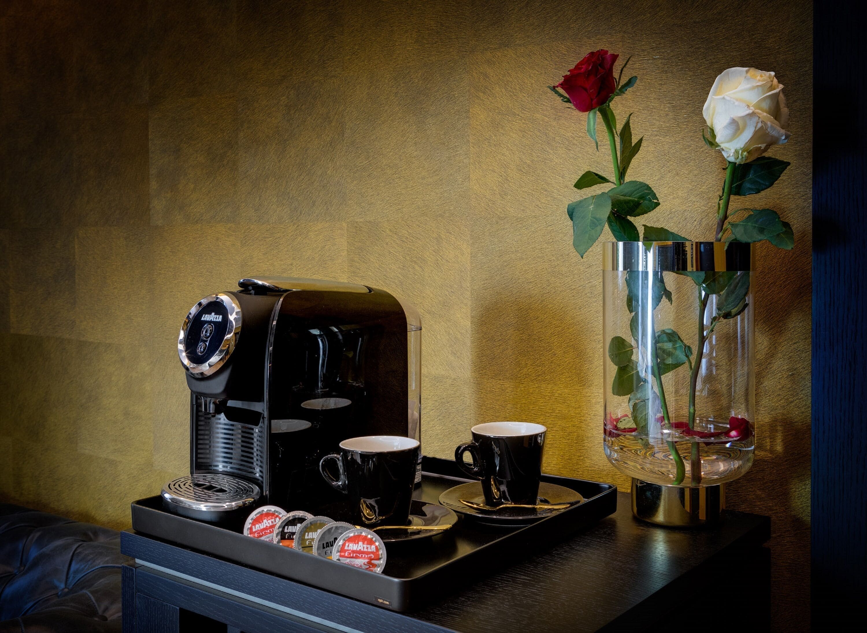 A coffee machine, a cup, and a vase with roses on a tray.