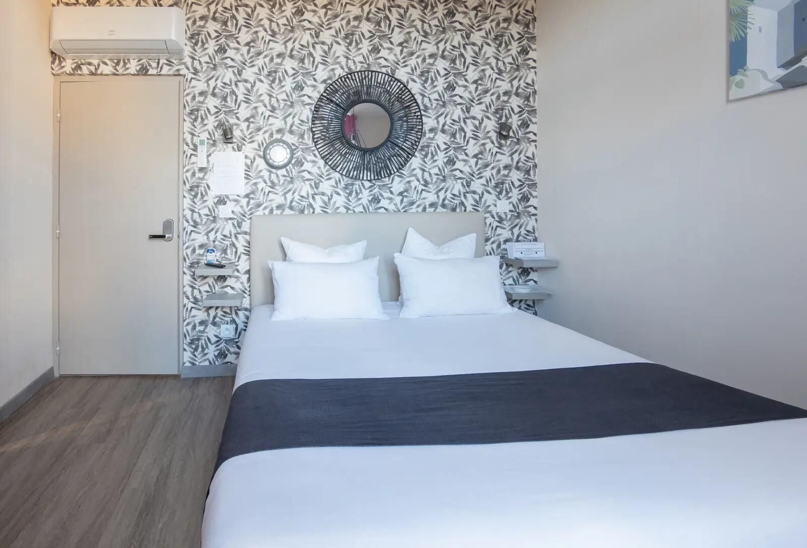 A neatly arranged hotel room with a bed, a wall-mounted mirror, and a patterned wallpaper.