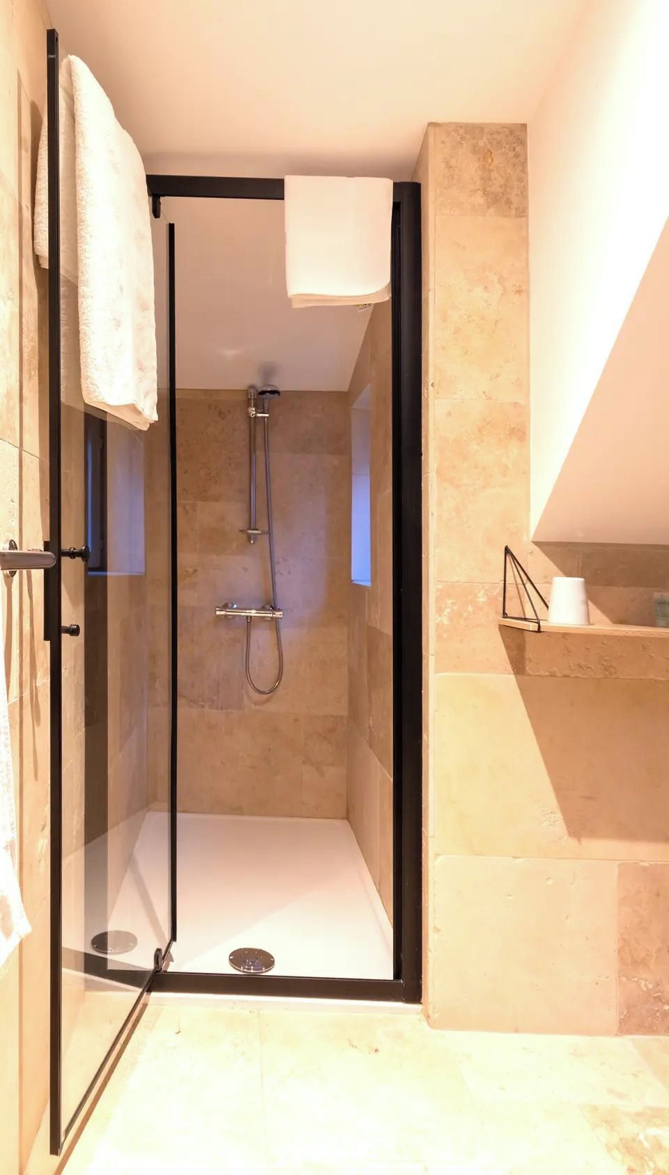 A glass-enclosed shower with a handheld showerhead, a towel rack, and a toilet paper holder on a beige tiled wall.