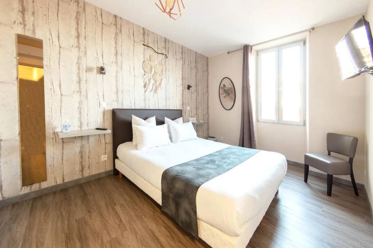 A hotel room with a large bed, a wooden accent wall, a small desk, a chair, and a window with curtains.