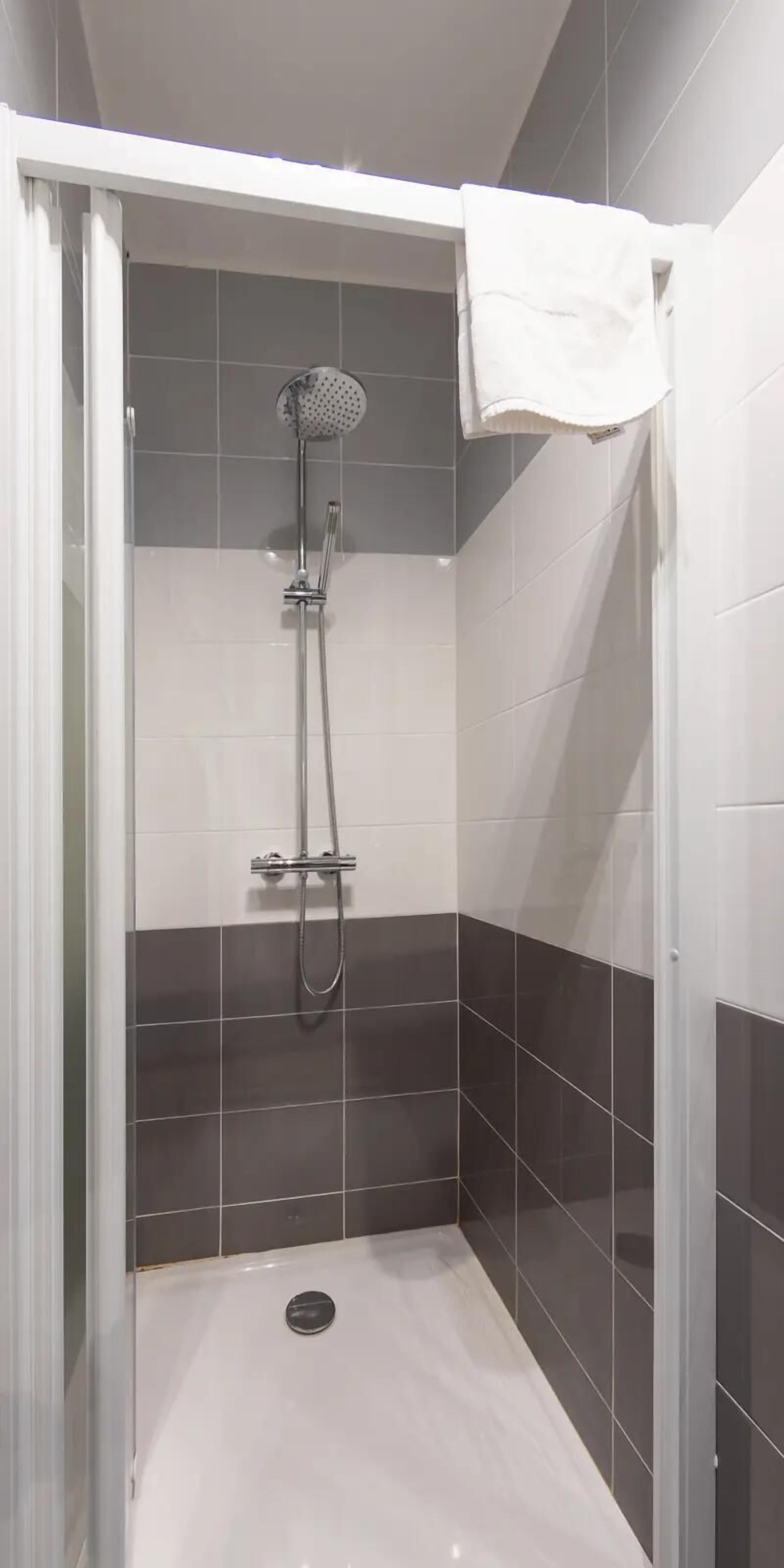 A shower with a handheld showerhead and a wall-mounted faucet, a white towel hanging on the wall, and a drain in the floor.