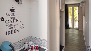 A modern kitchen with a coffee machine, cups, and a wall clock. A hallway leads to a door with a view of the outside. - Le Relais de la Poste (Gourdon)