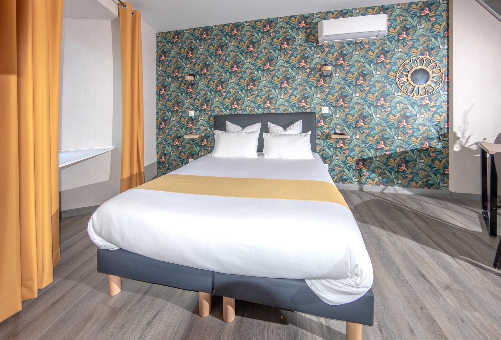 Un lit bien fait, recouvert d’une courtepointe blanche et bleue, un plancher en bois et un papier peint à motifs floraux.