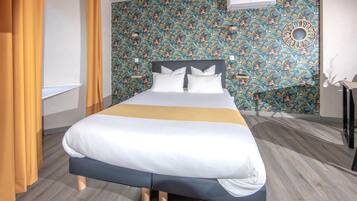 Un lit bien fait, recouvert d’une courtepointe blanche et bleue, un plancher en bois et un papier peint à motifs floraux.