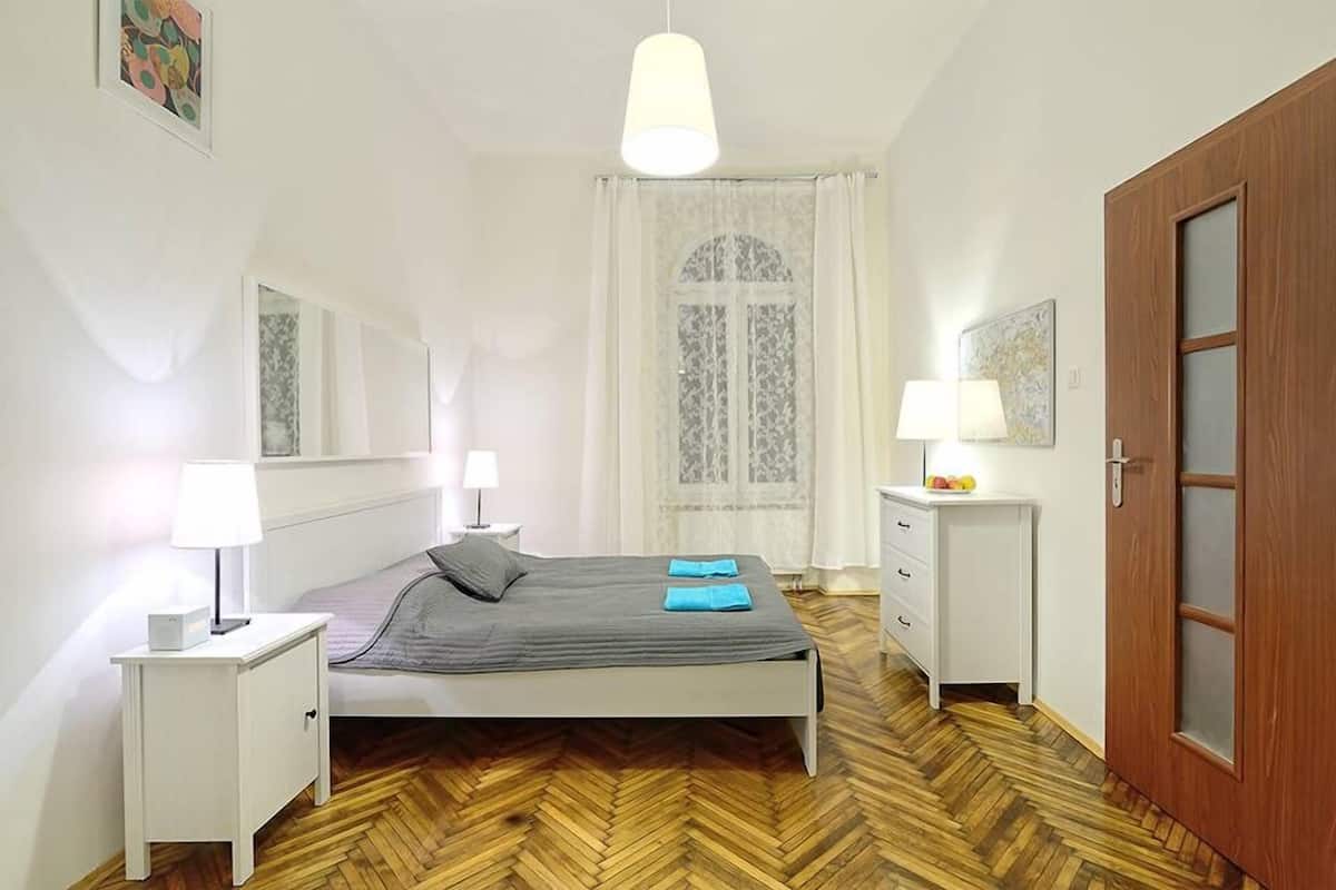 A bedroom with a bed, bedside tables, a wooden floor, a door, and a window with curtains.