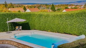 A rectangular swimming pool with a clear blue water surface, surrounded by a brick wall and a stone border. There are two lounge chairs and a single umbrella providing shade. The pool is set against a backdrop of a well-maintained hedge and a residential area with houses and trees in the distance.