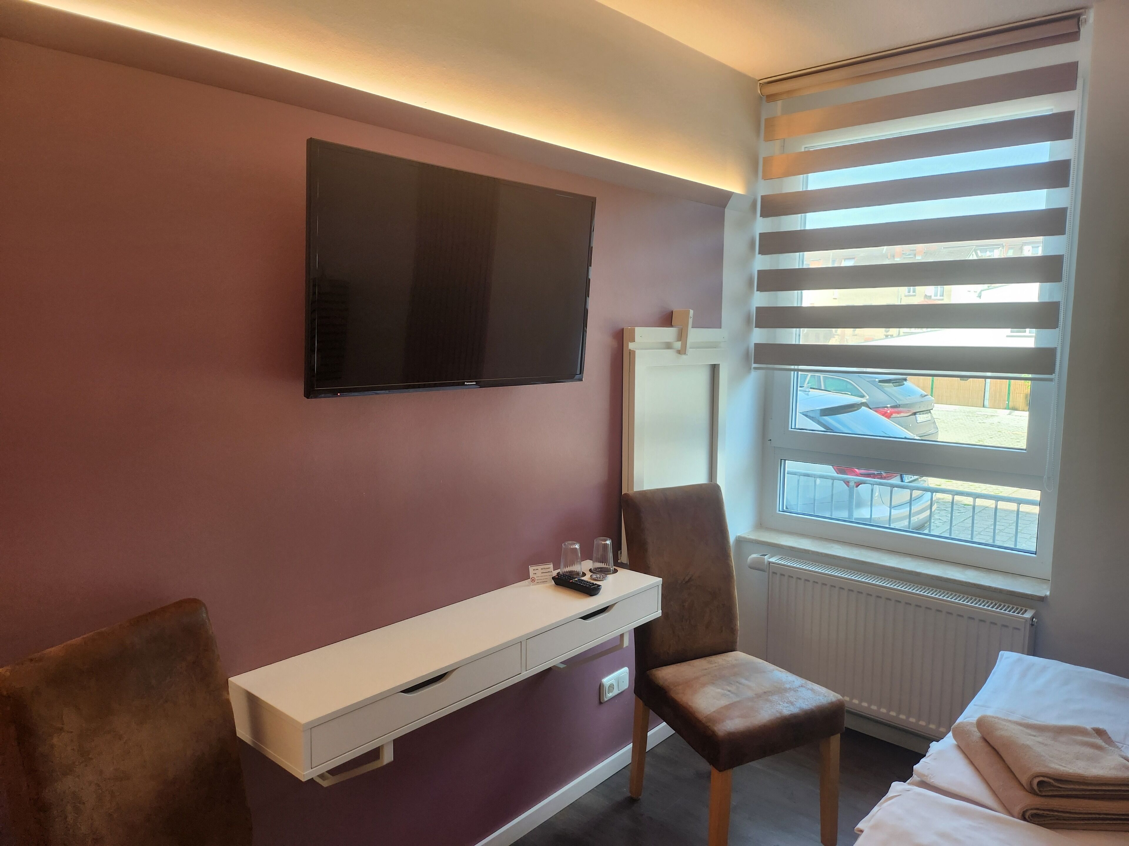 Une chambre d’hôtel équipée d’un téléviseur à écran plat fixé au mur, d’un bureau avec une chaise et d’une fenêtre avec des stores.