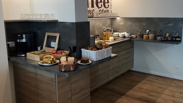 A modern kitchen with a coffee station, including a coffee machine, a variety of breads, fruits, and a sign for 'Cappuccino'. - Hotel Weinert (Neubrandenburg)