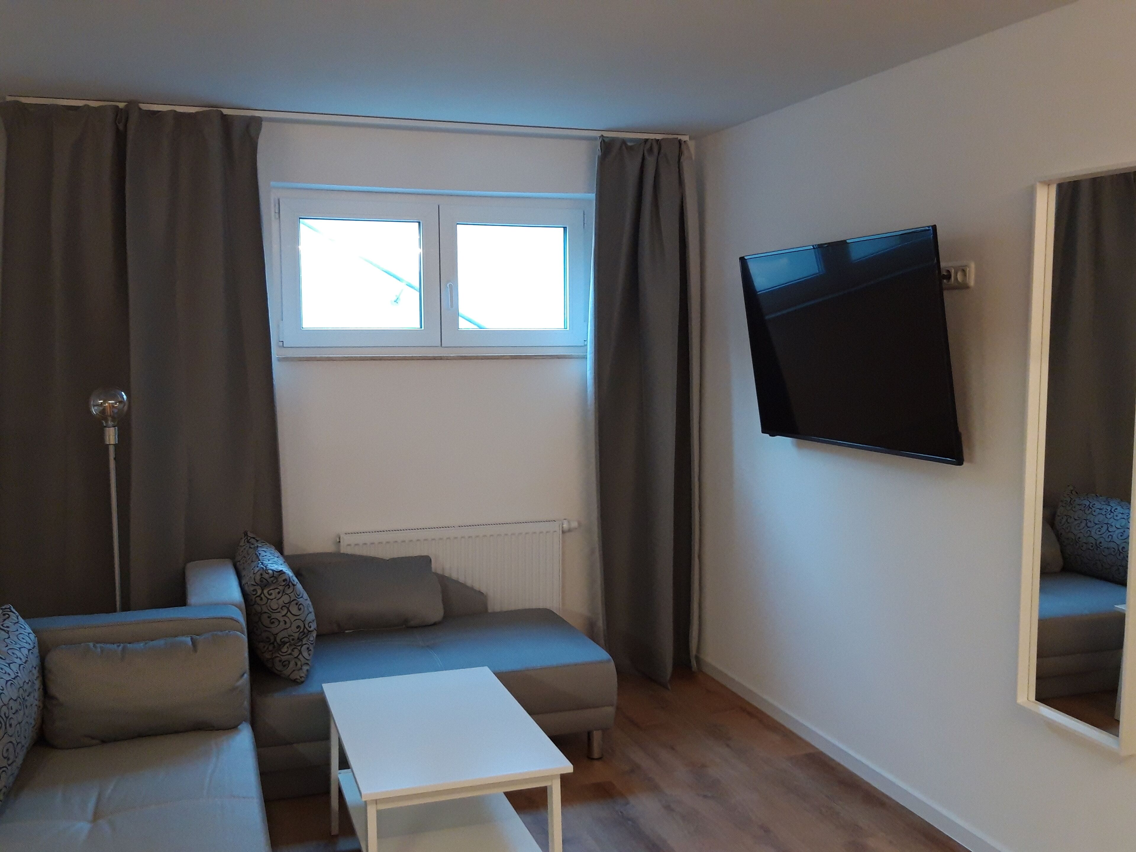 A living room with a sofa, a small table, a TV mounted on the wall, and a window with curtains.