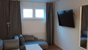 A living room with a sofa, a small table, a TV mounted on the wall, and a window with curtains.