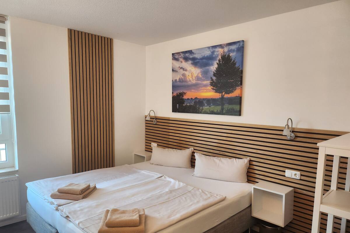A neatly made bed with two pillows and two folded towels, a bedside table, a wall-mounted lamp, and a painting of a sunset.