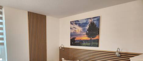 Ein ordentlich bezogenes Bett mit zwei Kissen und zwei gefalteten Handtüchern, ein Nachttisch, eine Wandlampe und ein Gemälde vom Sonnenuntergang.