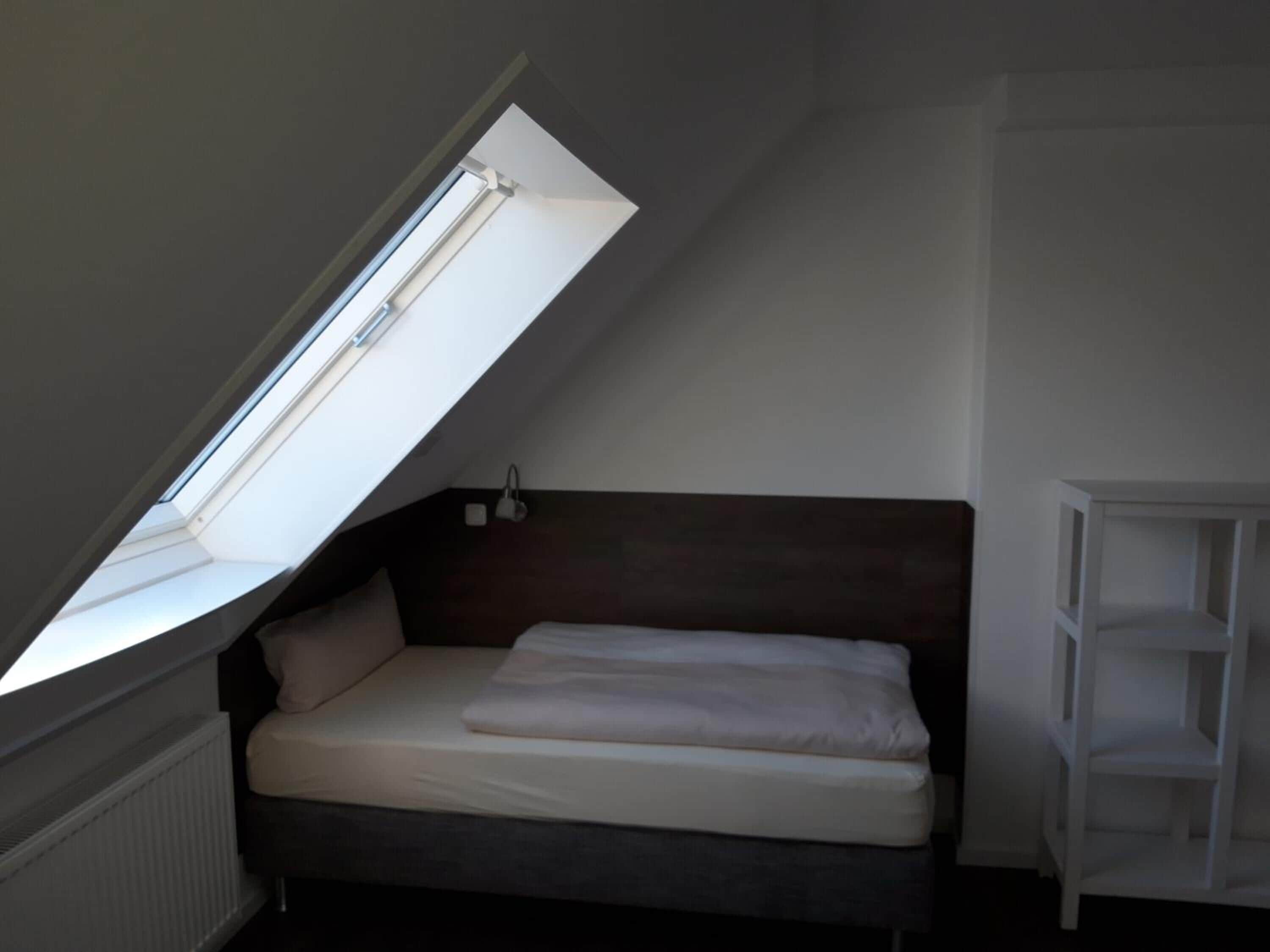 A small, dimly lit room with a slanted ceiling, a single bed, a white shelf unit, and a skylight.