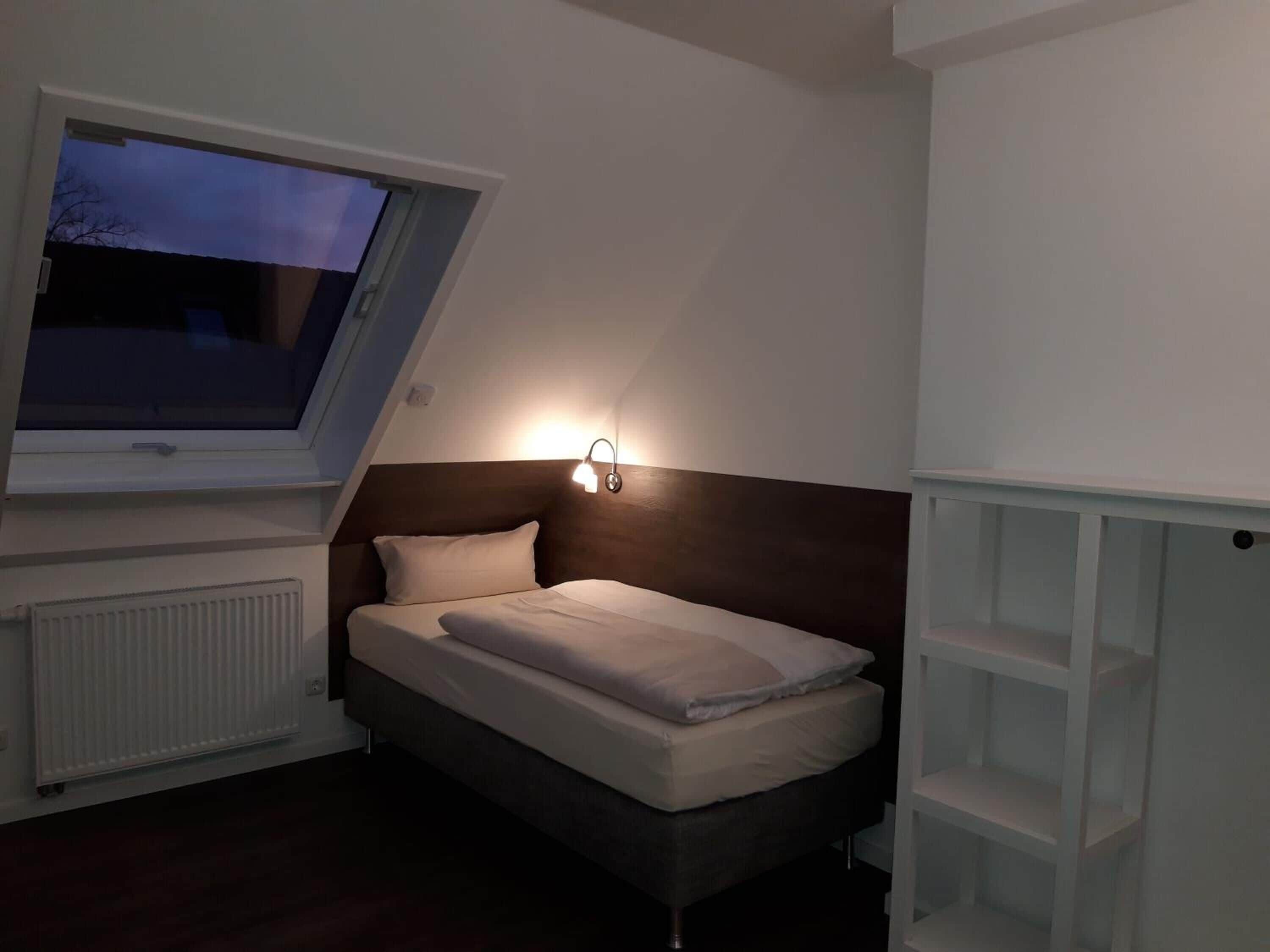 Un dormitorio con cama, mesita de noche, ventana y lámpara fijada a la pared.