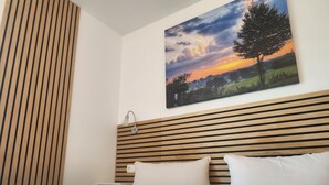 A bedroom with a wooden slat headboard, a painting of a tree and sunset, and a neatly made bed with pillows. - Hotel Weinert (Neubrandenburg)