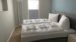 Een net opgemaakt bed met witte lakens en kussens, een radiator, een raam met zonwering, en een blauwe muur.