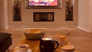 Een gezellige woonkamer met een open haard, een televisie bevestigd aan een stenen muur, een bijzettafeltje met kopjes en een theepot, en een afstandsbediening.