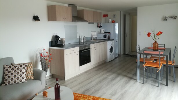 A modern kitchen with a built-in oven, microwave, and washing machine. There is a dining area with a table and chairs, and a living room with a sofa and a coffee table.