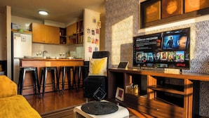 Sala de estar com televisão exibindo um filme, área de bar com banquetas, cozinha ao fundo e parede com padrão.