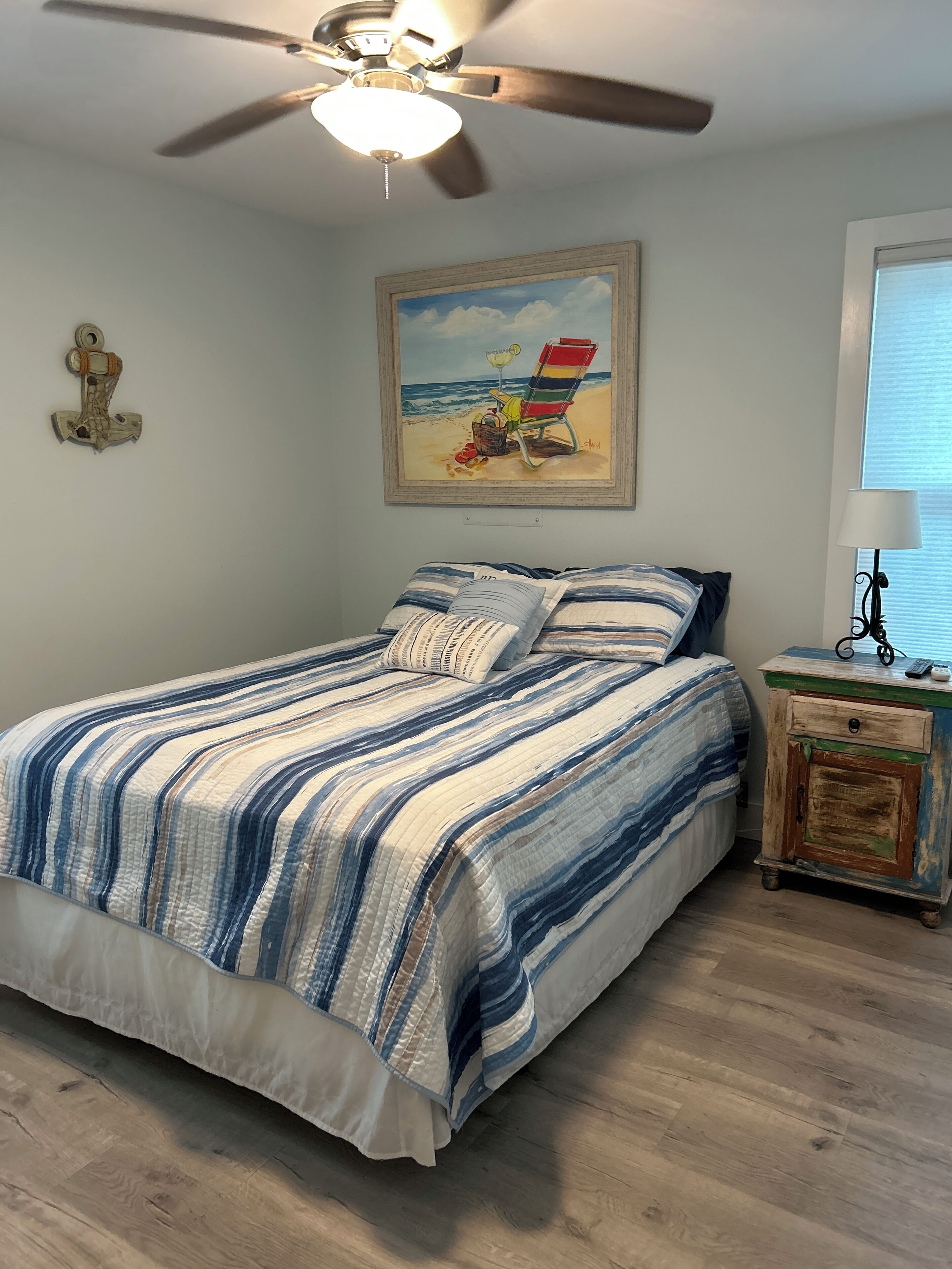 A bedroom with a bed, a ceiling fan, a painting of a beach scene, a nightstand, and a window with blinds.