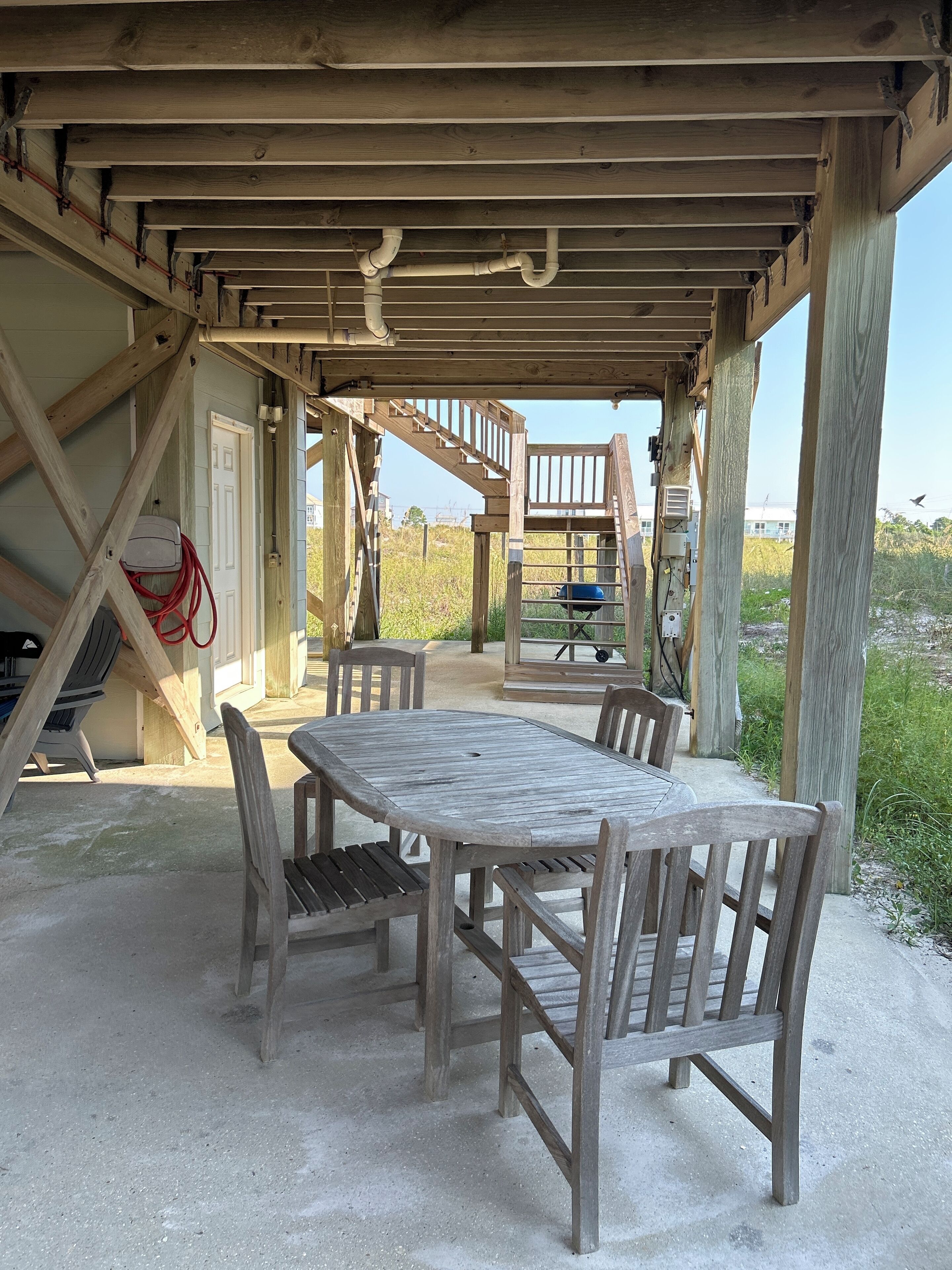 Un espace extérieur abrité, doté d’une table et de chaises en bois, d’un barbecue et d’un escalier menant à un niveau supérieur.