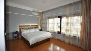 A bedroom with a wooden bed, a nightstand, a wall-mounted air conditioner, and a window with sheer curtains. - Line-Residence (Douala)