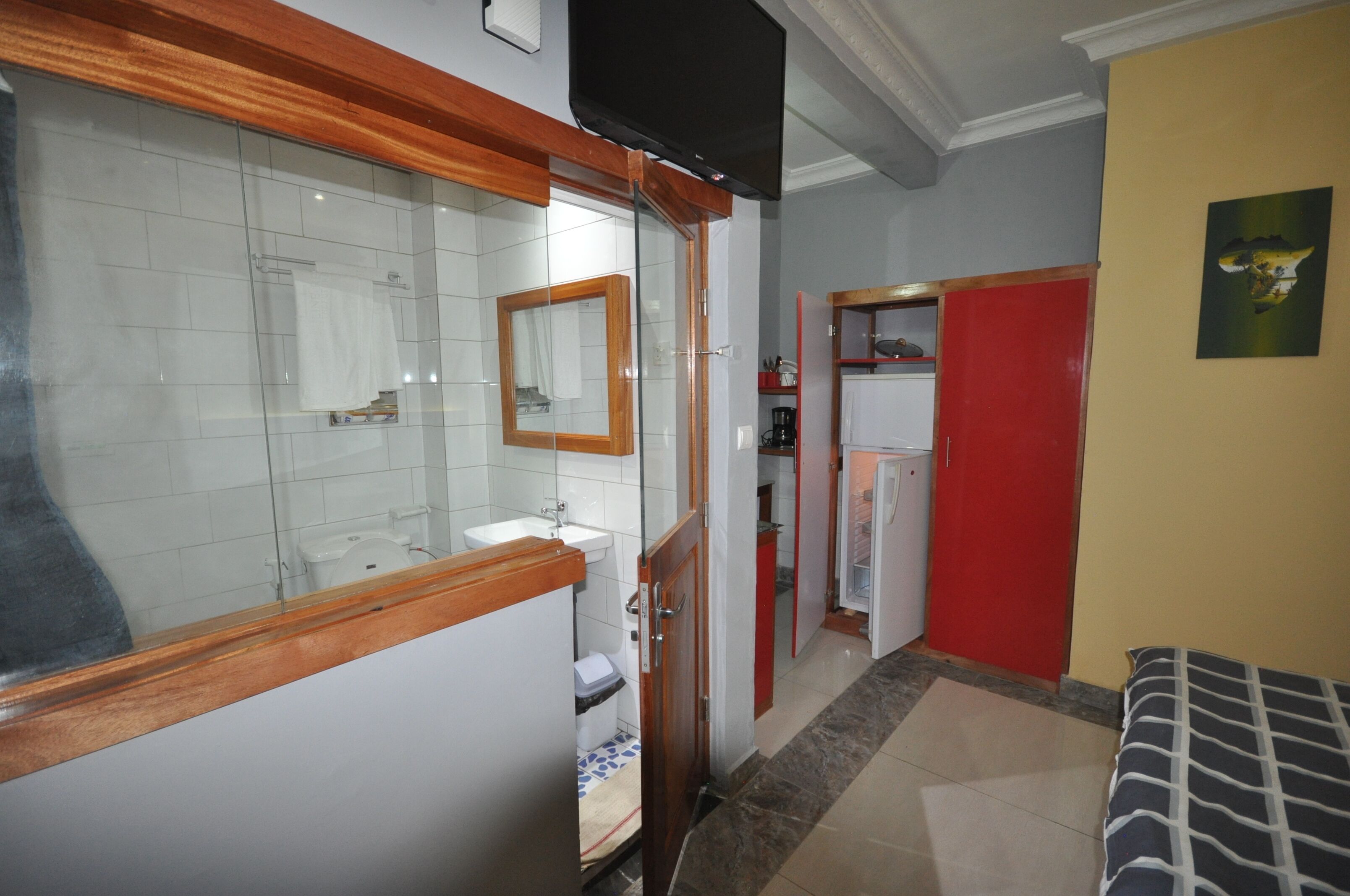 Un bagno con box doccia in vetro, un lavandino e un water. Si nota un'armadio rosso e un letto con coperta a righe.
