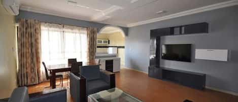 A modern living room with a dining area, a kitchen in the background, and a flat-screen TV mounted on the wall.