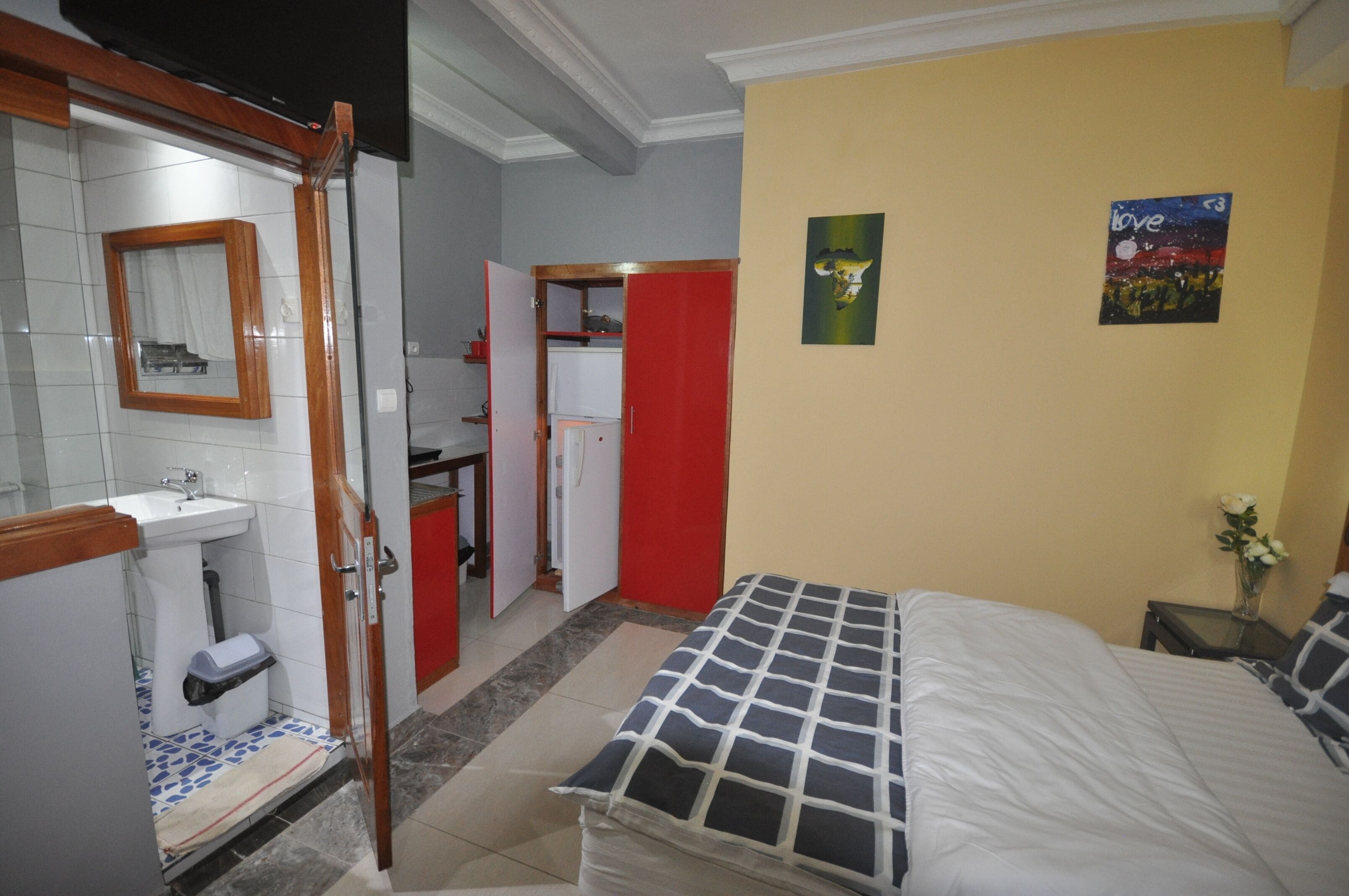 A bedroom with a bed, a red wardrobe, a bathroom with a sink, and a mirror.