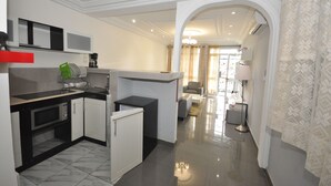 A modern kitchen with white cabinetry, a black countertop, and a microwave. There is a small refrigerator and a sink with a faucet. The kitchen leads to a living area with a sofa and a lamp. - Line-Residence (Douala)