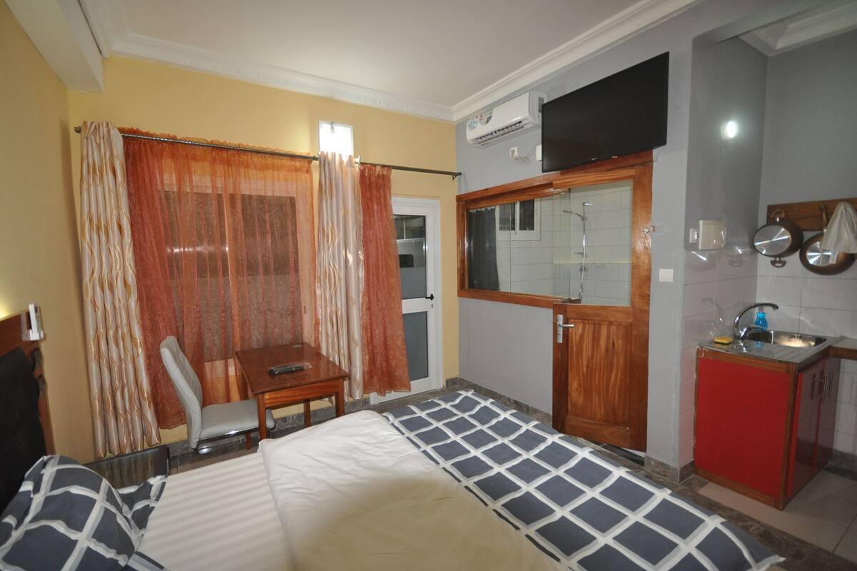 A hotel room with a bed, a desk, a chair, a small kitchenette, and a bathroom with a shower.