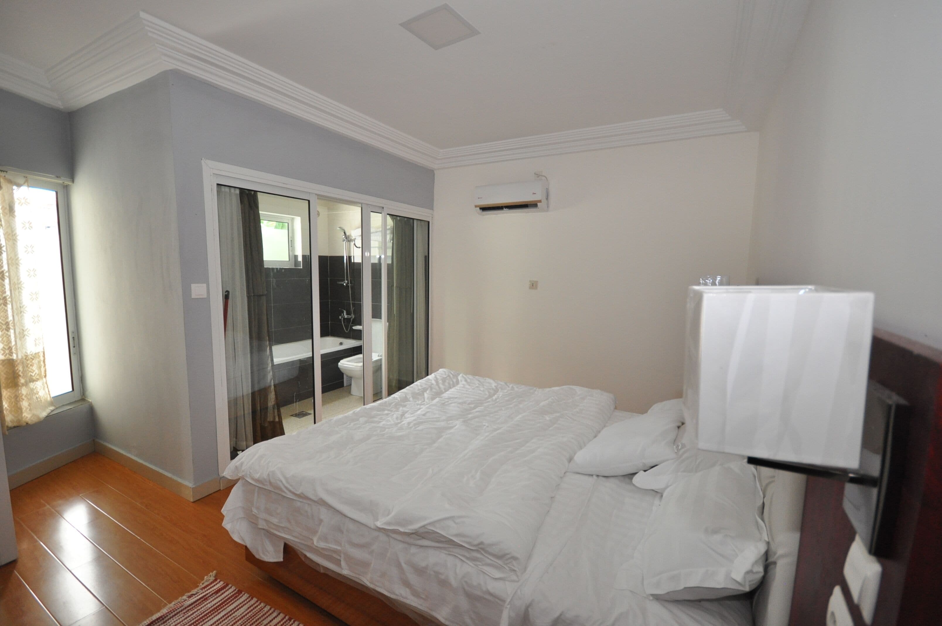 A bedroom with a bed, a sliding glass door to a bathroom, and a white air conditioning unit on the wall.