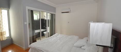 A bedroom with a bed, a sliding glass door to a bathroom, and a white air conditioning unit on the wall.