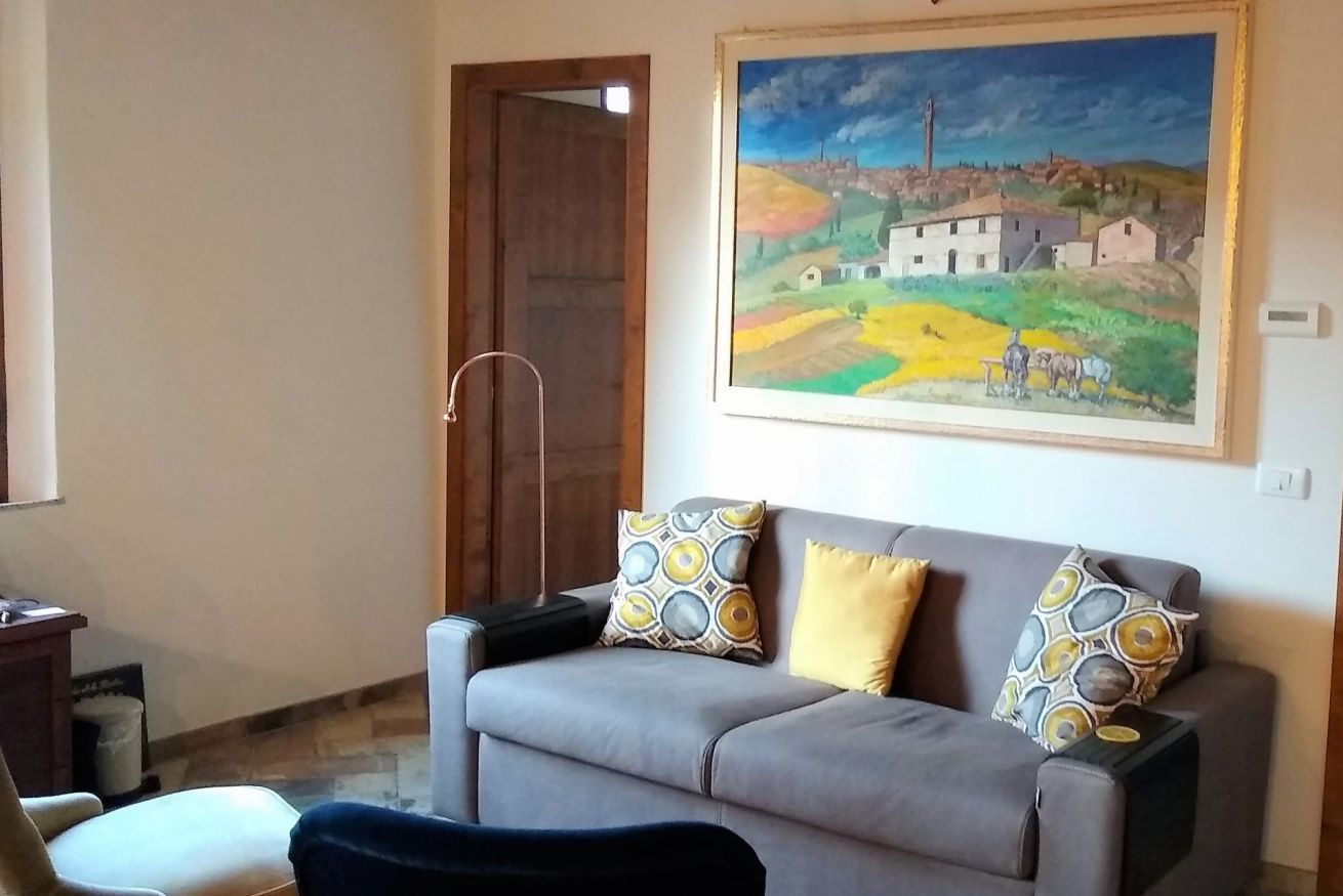 Un salon avec un canapé gris, deux coussins à motifs, un tableau représentant un paysage rural et une porte en bois.