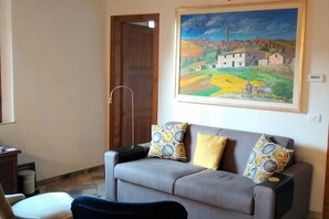A living room with a grey sofa, two patterned cushions, a painting of a rural landscape, and a wooden door.
