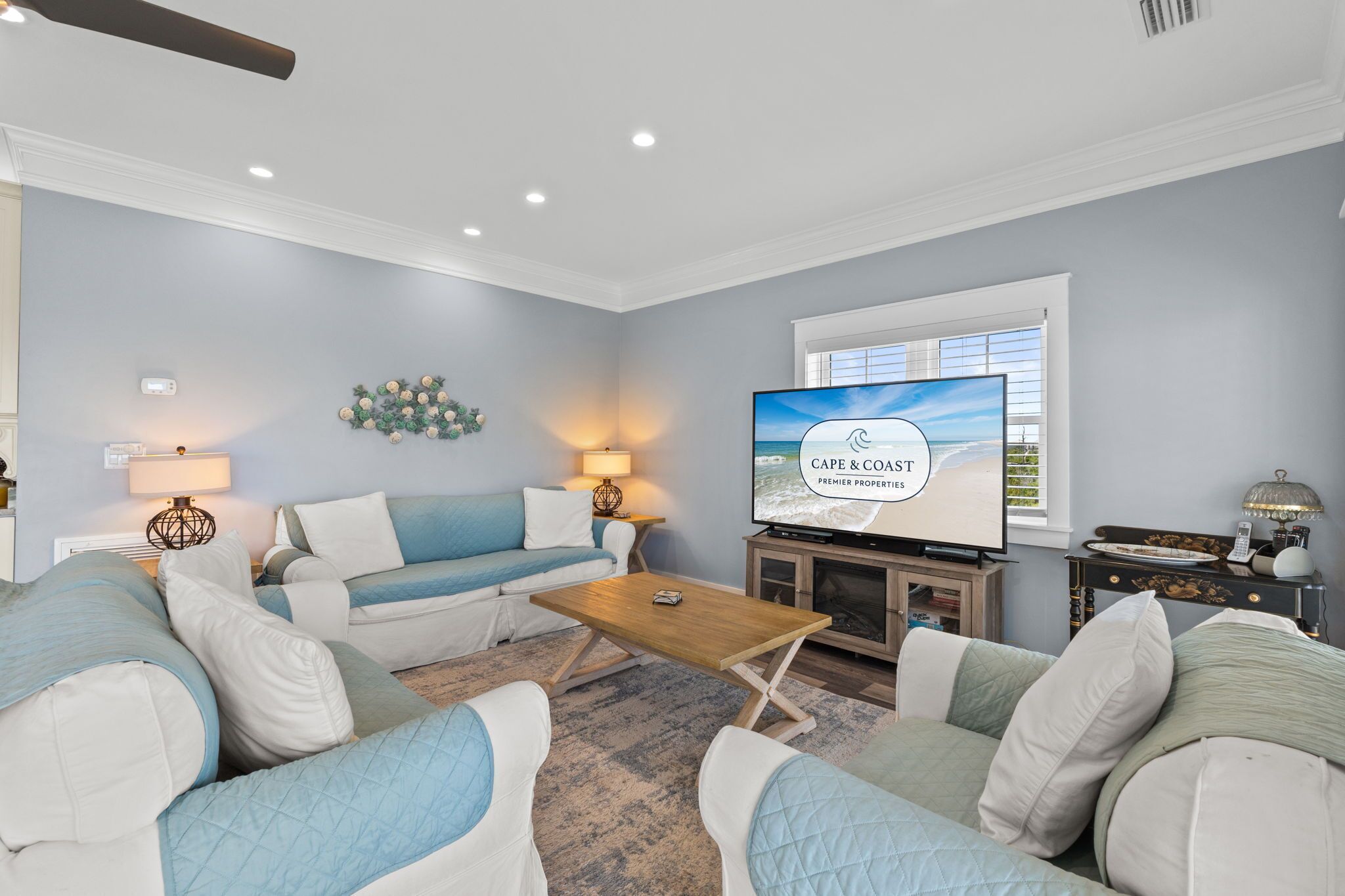 Un salon comprenant un canapé, une table basse, un téléviseur diffusant l'émission « Cape & Coast Premier Properties », et une fenêtre équipée de stores.