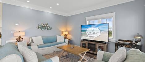 Un salon comprenant un canapé, une table basse, un téléviseur diffusant l'émission « Cape & Coast Premier Properties », et une fenêtre équipée de stores.