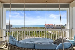 Een balkon met een rieten schommelstoel, uitzicht op de oceaan, en een gebouw op de achtergrond.